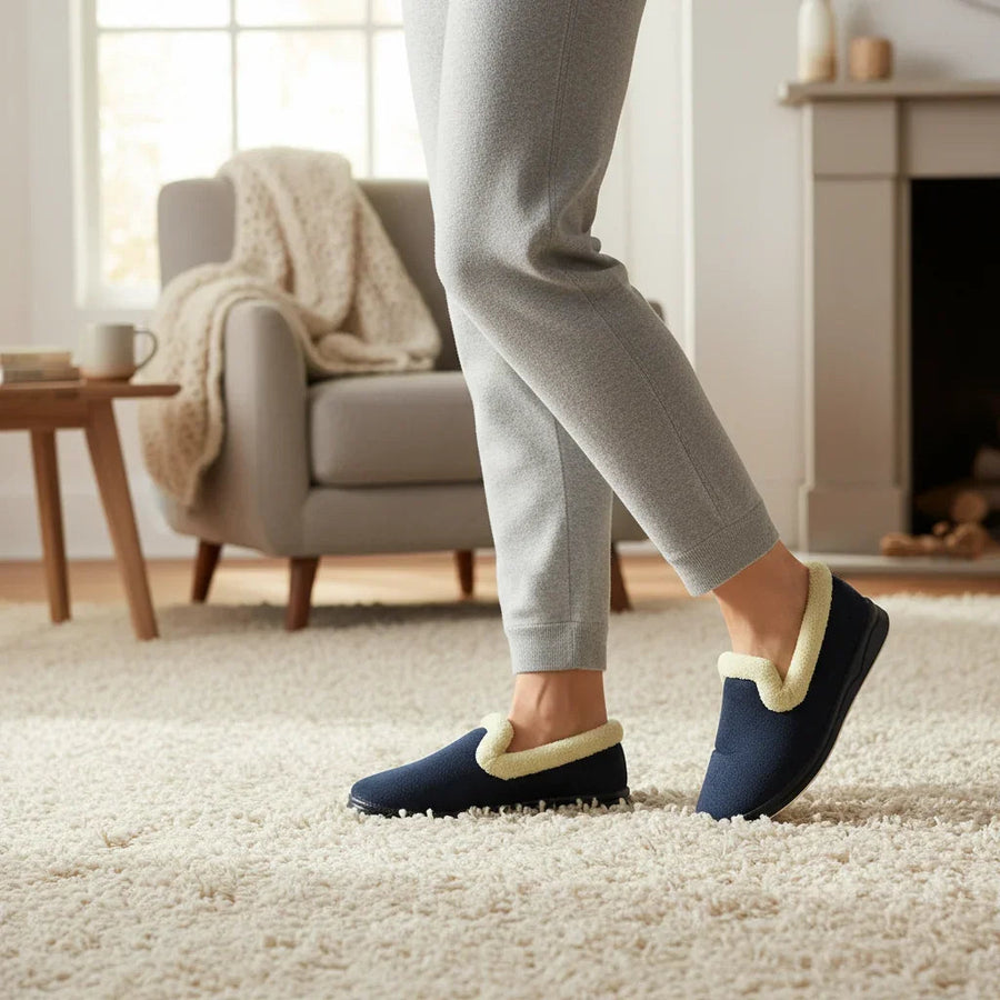 Navy plush slip-on slippers with light tan paw appliqué, worn with gray pants
